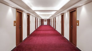 Ein langer, schmaler Hotelgang mit rotem Teppich, Holztüren und eingelassener Beleuchtung an der Decke.