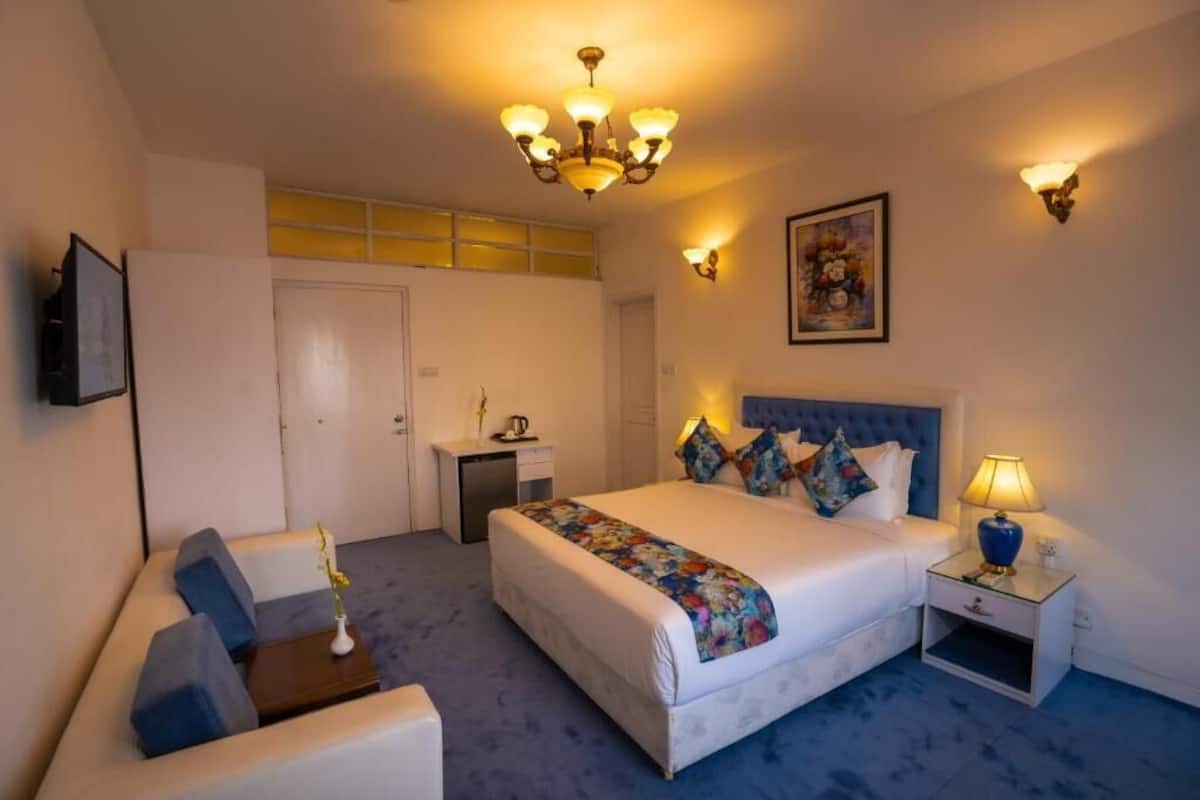 Ein Hotelzimmer mit einem großen Bett, einem Nachttisch mit Lampe, einem kleinen Schreibtisch, einem Sessel und einem an der Wand montierten Fernseher.