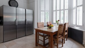 A kitchen with a stainless steel refrigerator, a wooden dining table with chairs, and a window with white shutters. - EBS London Stay (London)