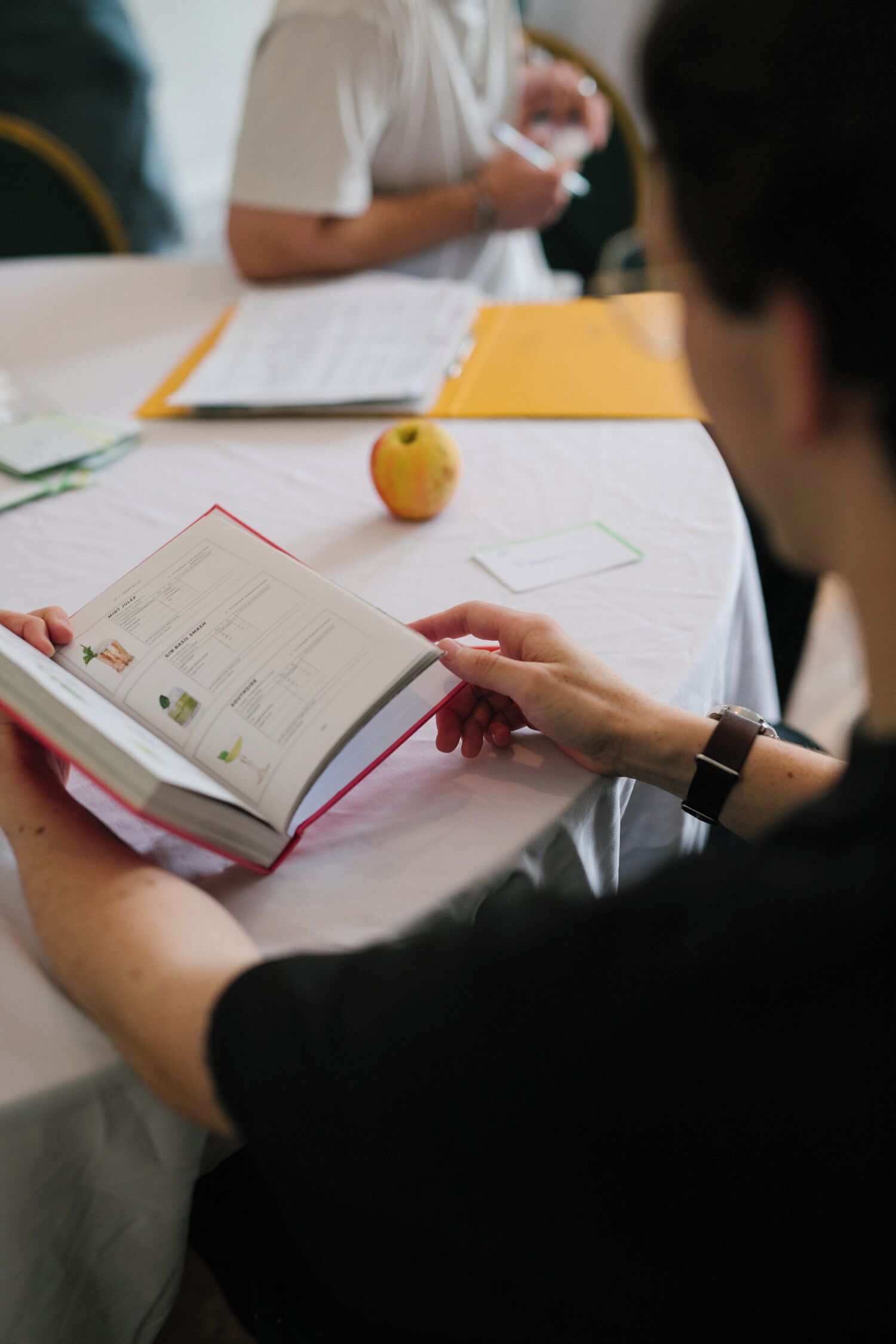 A person reading a book at a table with a yellow folder and a green pen.