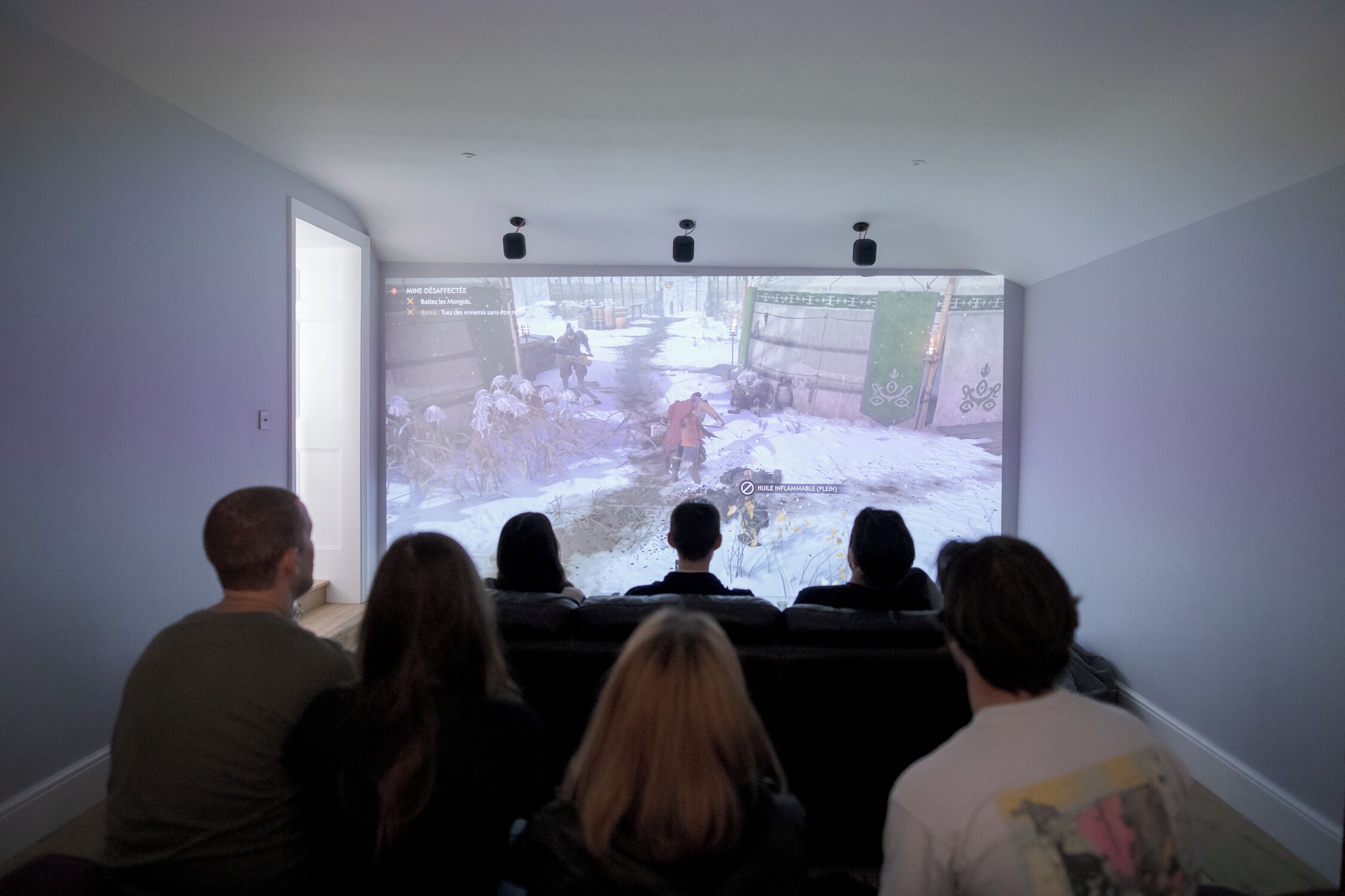 Un grupo de personas observando un video proyectado en una sala con una pantalla grande, una puerta y un techo con altavoces instalados.