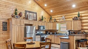 A cozy log cabin interior with wooden walls and ceilings, a central kitchen area with modern appliances, a dining table with chairs, and a small bar area. - Two Bears Cabin (Cabin 162) (Eagar)