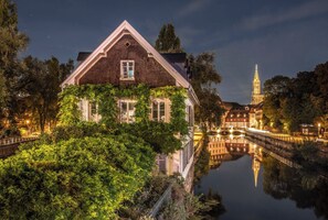 Ein historisches Gebäude mit einem markanten Turm, umgeben von üppigem Grün und einem ruhigen Wasserspiegel, der die Architektur widerspiegelt.