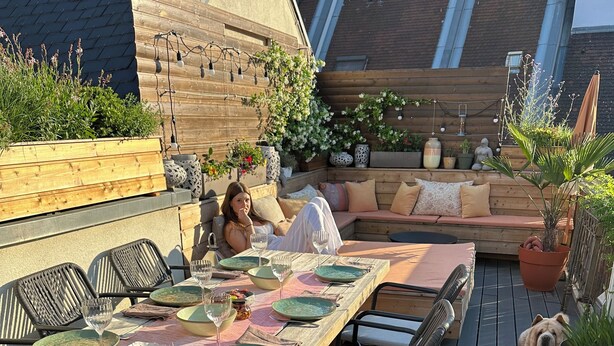 Un espacio de comedor al aire libre con una mesa de madera larga, múltiples sillas y una zona de estar con cojines.