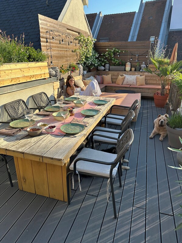 An outdoor dining area with a long wooden table, multiple chairs, and a cushioned seating area.