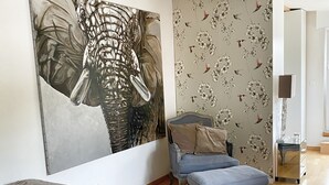 Un dormitorio con un cuadro abstracto grande, papel tapiz floral, una cama con colcha gris, un sillón azul y suelo de madera.