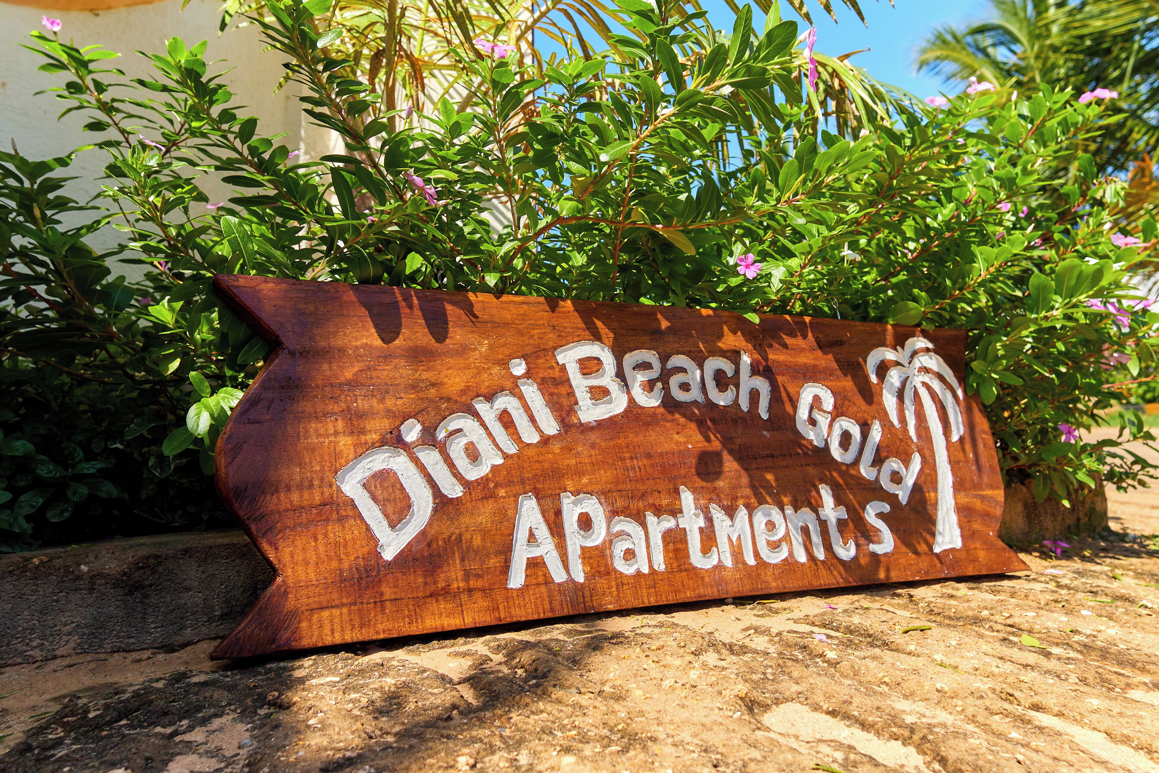 Une enseigne en bois indiquant « Diani Beach Gold Apartments », avec un symbole représentant un palmier.