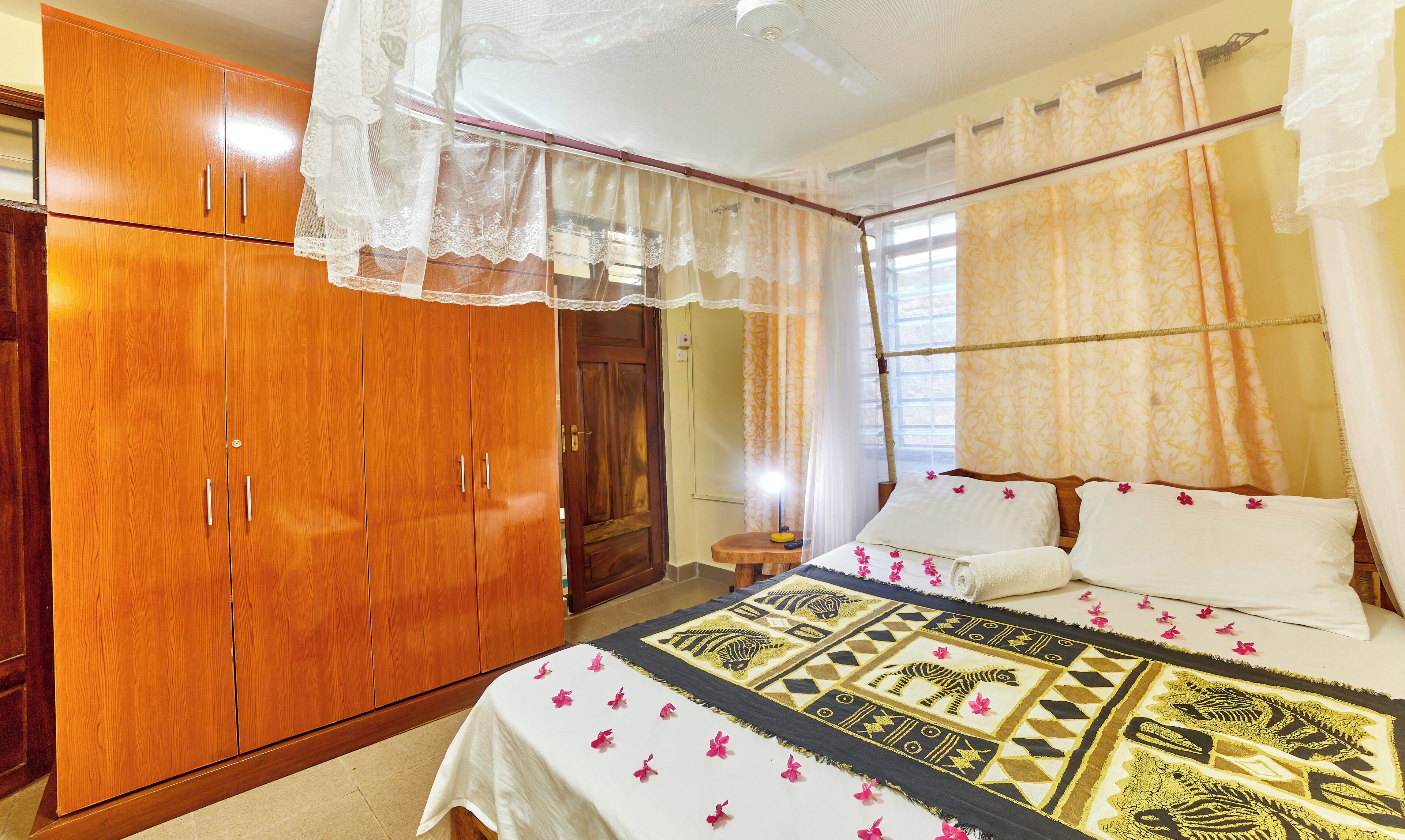 A bedroom with a wooden wardrobe, a bed with a patterned bedspread, and sheer curtains.