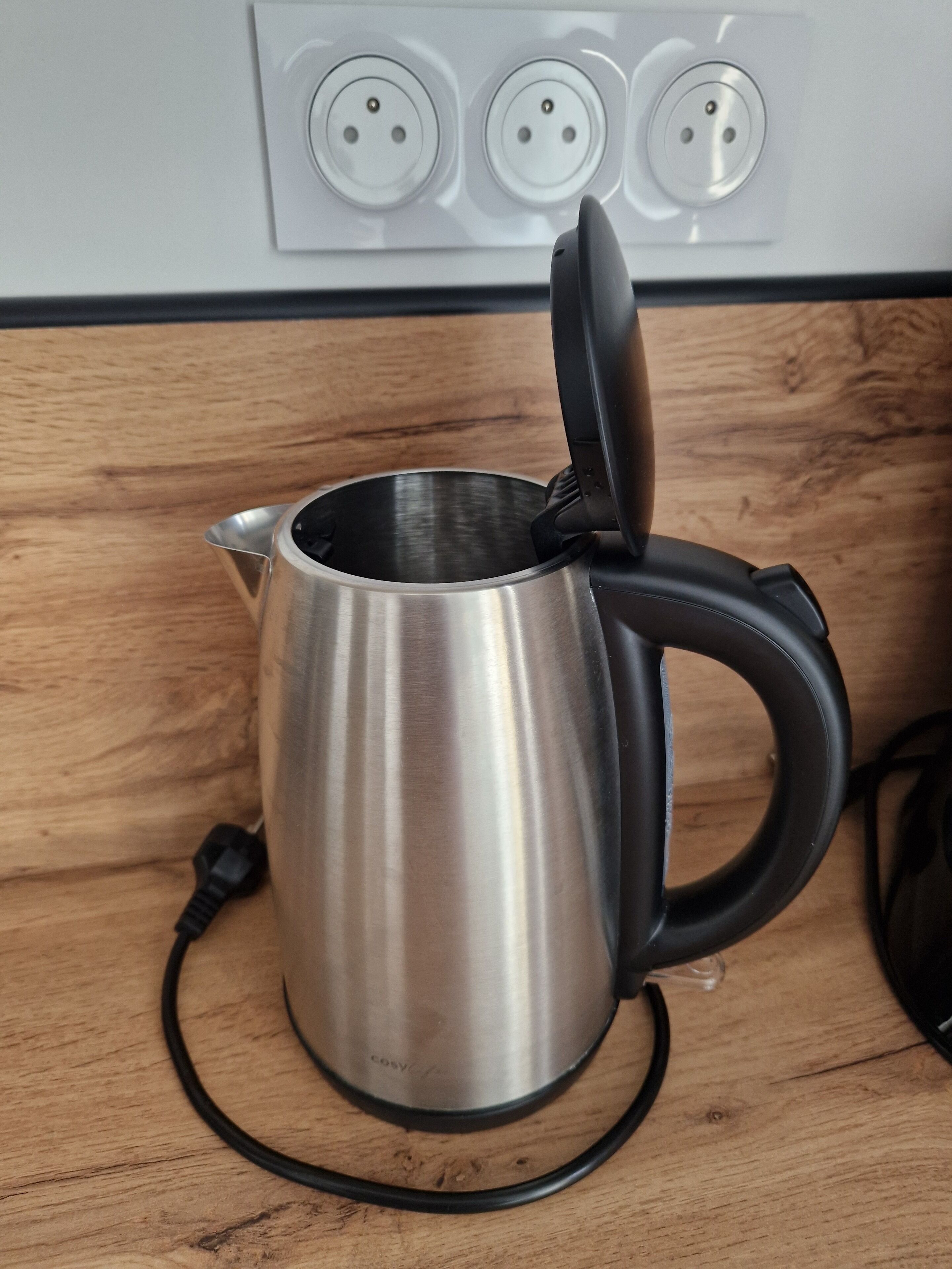 Een roestvrijstalen elektrische waterkoker met een zwart handvat en snoer op een houten aanrecht.