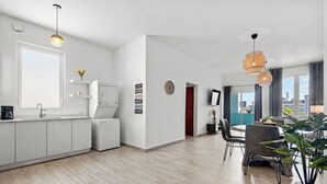 Et moderne køkken med hvide skabe, en indbygget ovn og et stort vindue. Der er et spiseområde med et bord og stole, samt en stue med en sofa og en plante.