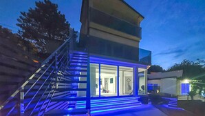 A modern two-story building with a glass facade, blue lighting accents, and a visible interior with a wheelchair. - Villa With Swimming Pool (Matulji)
