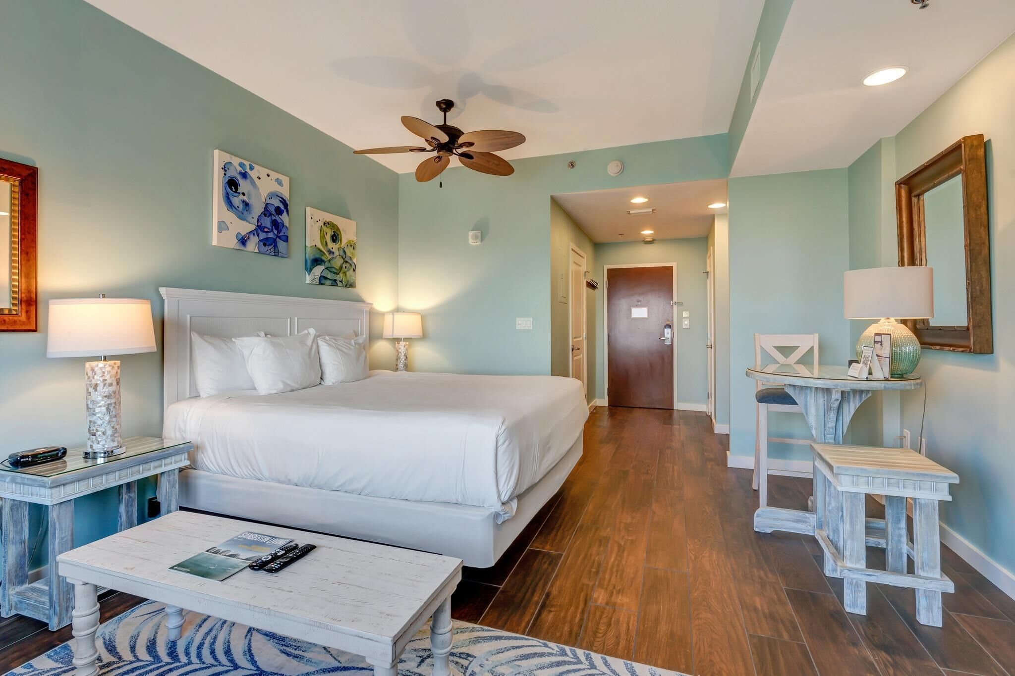 A bedroom with a large bed, two bedside tables with lamps, a ceiling fan, and artwork on the walls.