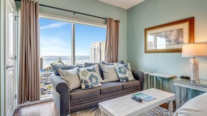 A living room with a sofa, a coffee table, a lamp, and a view of the city through large windows. - Serene Gulf Views: 2-Bedroom 2-Bathroom Retreat at Luau, Panoramic Gulf Views at Sandestin Golf and Beach Resort! (Miramar Beach)