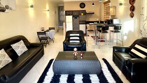 Un salon moderne avec un canapé en cuir noir, un tapis à rayures et une table basse. On aperçoit à l’arrière-plan un coin cuisine avec des tabourets de bar et une table à manger.