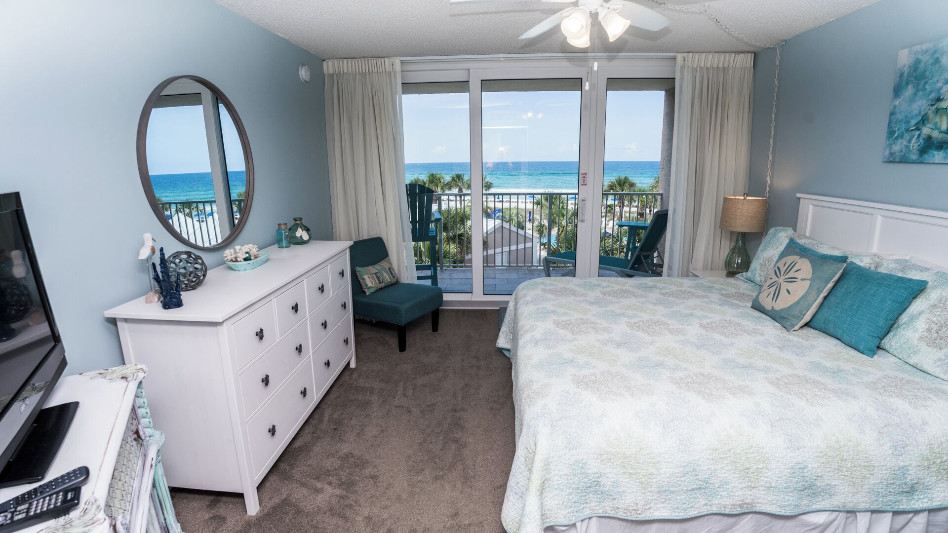 Een slaapkamer met een groot bed, een dressoir, een stoel, een televisie en uitzicht op zee vanaf een balkon.