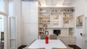 Une pièce avec une table blanche, quatre chaises, un vase rouge, une bibliothèque remplie de livres, des tableaux encadrés et un téléviseur.