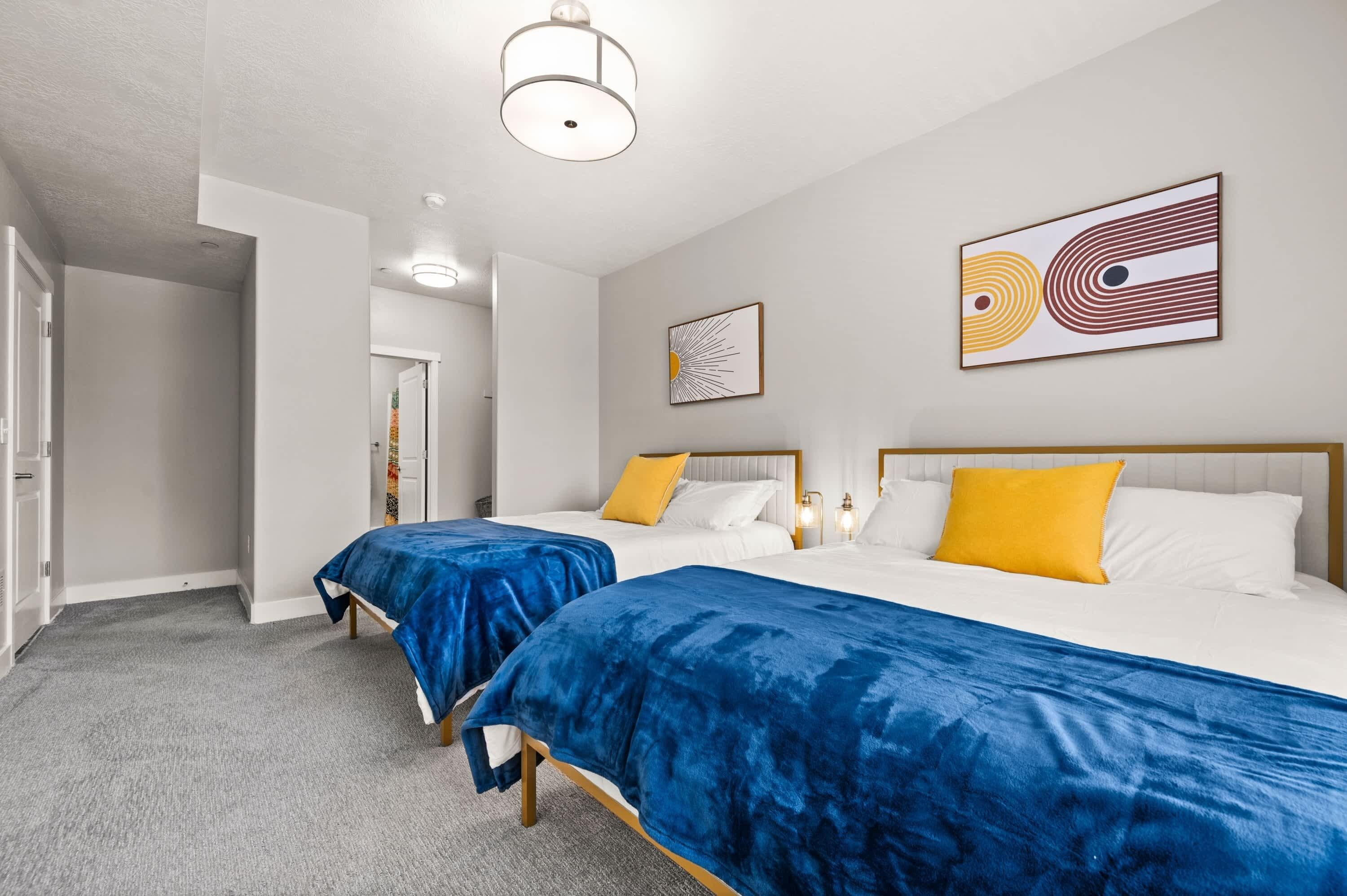 A hotel room with two beds, each with a blue bedspread and a yellow pillow. There are two wall-mounted artworks, a ceiling light, and a door leading to another room.