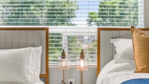 A hotel room with two beds, a round wooden table with two lamps, and a window with blinds.