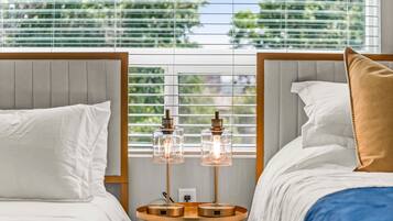 A hotel room with two beds, a round wooden table with two lamps, and a window with blinds.