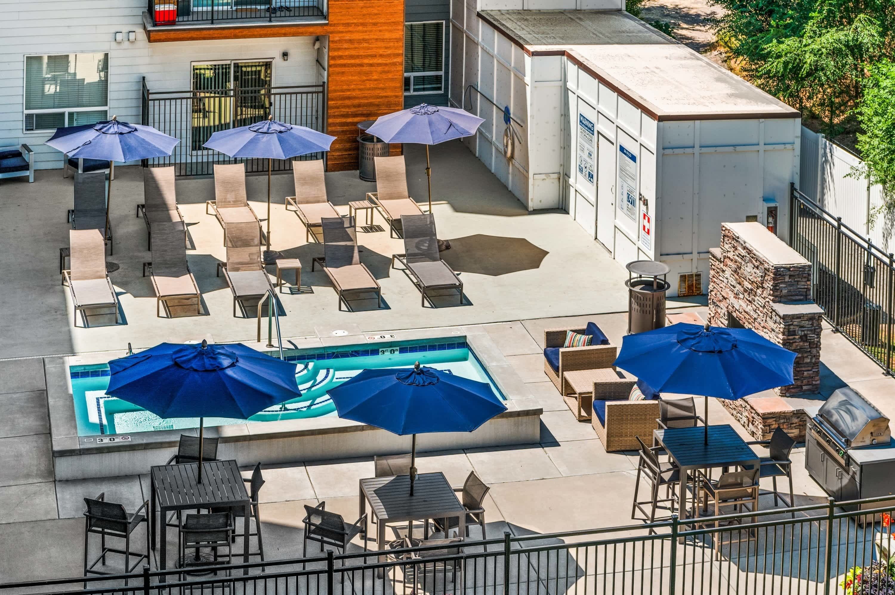 A rooftop pool area with lounge chairs and umbrellas, a barbecue grill, and a stone wall.