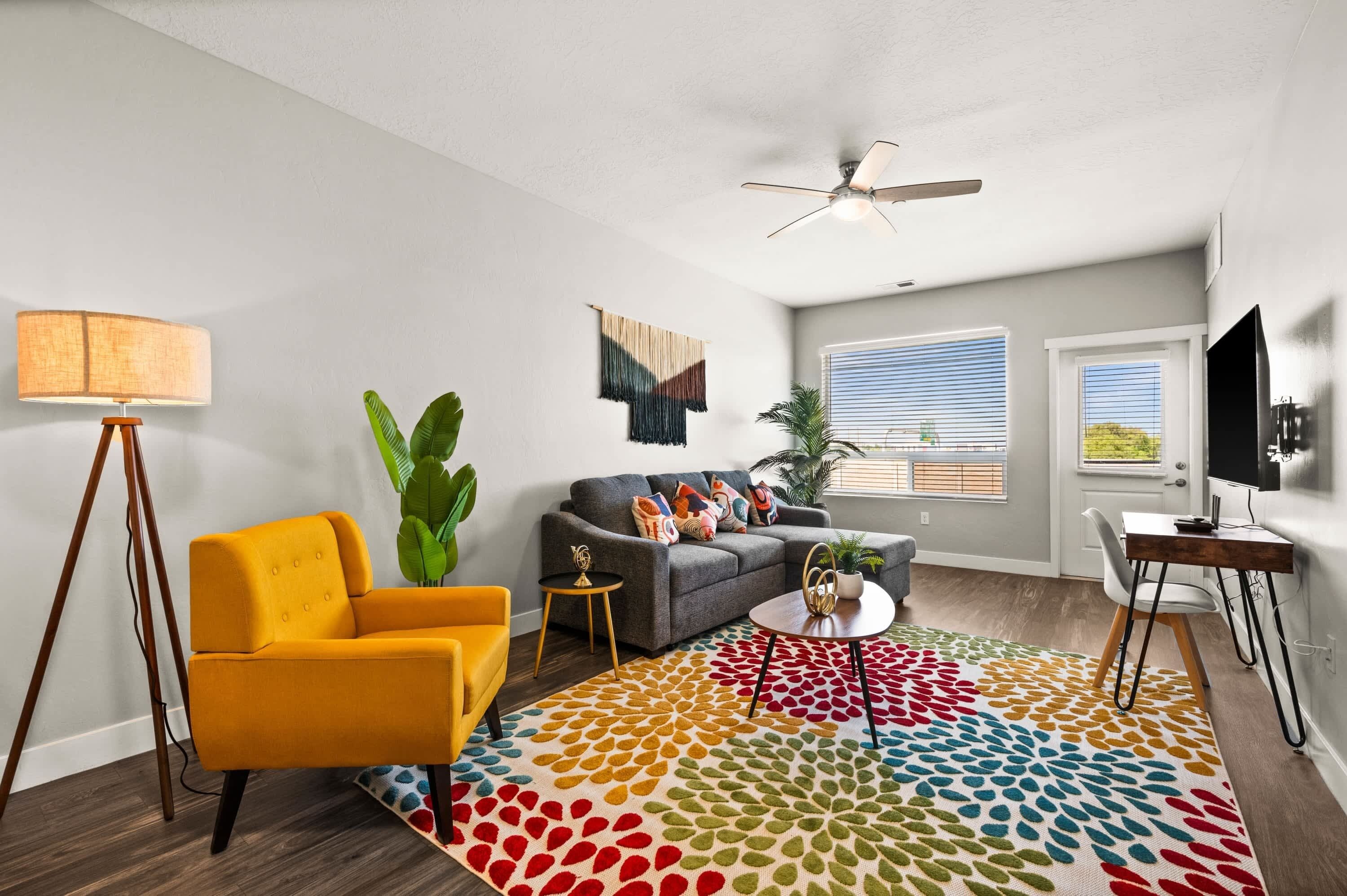 Una sala de estar moderna con sofá, sillón y mesa de centro, un ventilador de techo y un ventanal con persianas.