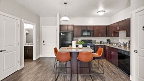 Une cuisine moderne avec des meubles en bois foncé, un réfrigérateur en acier inoxydable, un four à micro-ondes et un lave-vaisselle. Elle est dotée d’un plan de travail en granit et de deux tabourets de bar orange.