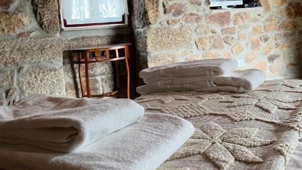 A stone-walled room with a bed, a television, and a window with curtains. - Casa de Campo Benfeito (Castro Daire)