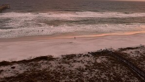 Une plage avec une jetée en bois qui s’avance dans l’océan, des vagues qui se brisent doucement sur le rivage et un ciel dégagé au-dessus.