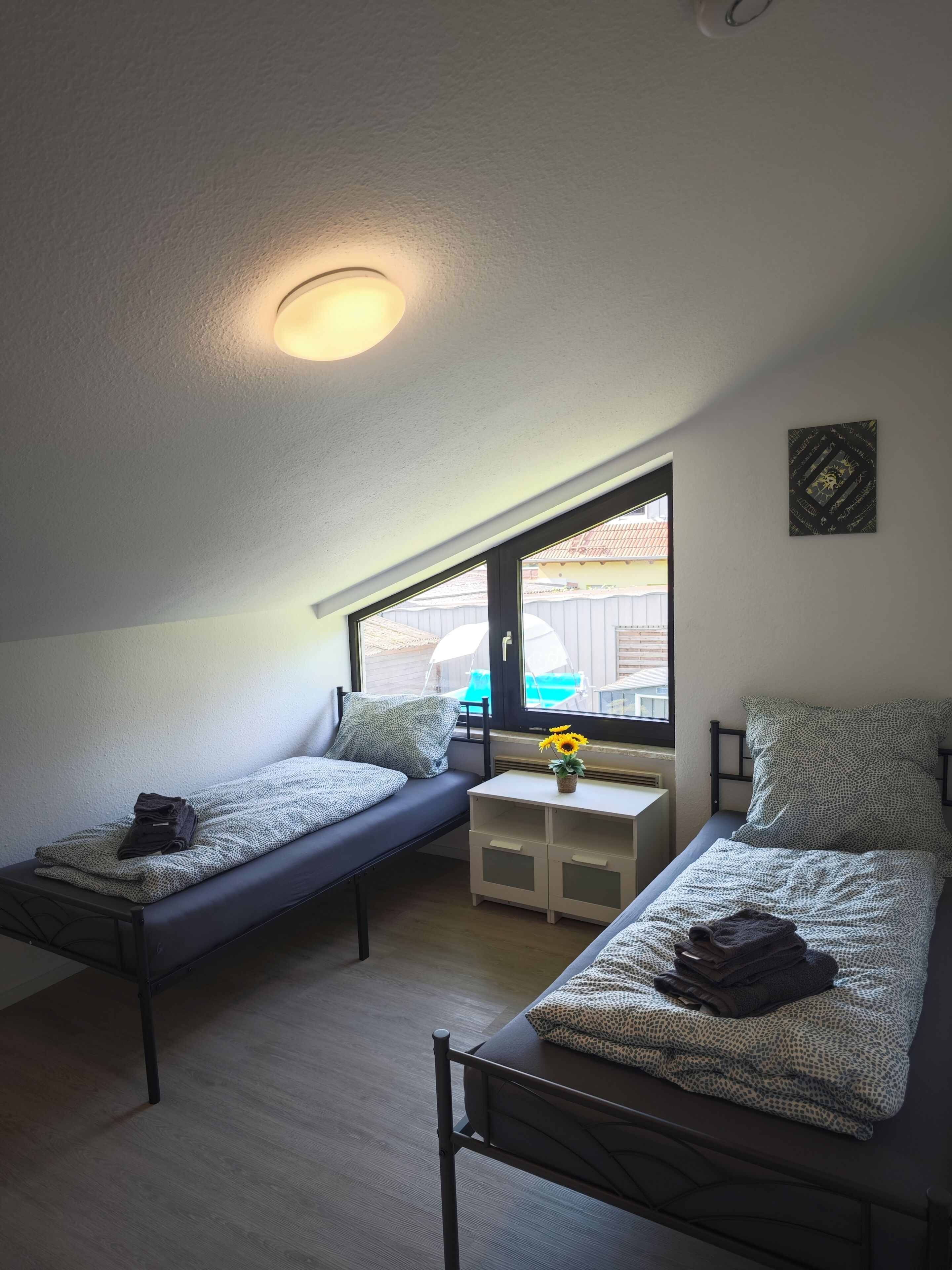 Ein kleiner, gut beleuchteter Raum mit zwei Einzelbetten, einem kleinen Tisch mit Blumenvase und einem Fenster mit Blick nach draußen.
