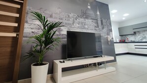 Un salon moderne avec un téléviseur à écran plat posé sur un meuble blanc, une grande plante en pot et une fresque représentant un paysage urbain sur le mur.