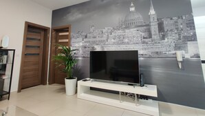 Un salon avec un grand téléviseur à écran plat posé sur un meuble blanc, une plante en pot et une porte en bois.