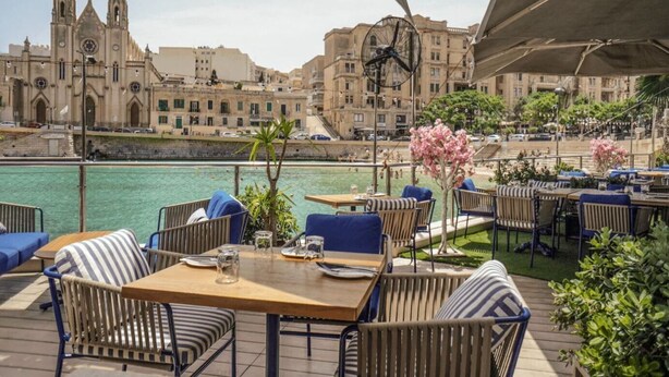 Un espace de restauration en plein air, avec des tables et des chaises, offrant une vue sur un plan d’eau et des bâtiments historiques.