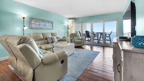 Una sala de estar con un sillón reclinable, un sofá, una mesa de centro y un mueble para televisión. Cuenta con un balcón con una mesa y sillas.