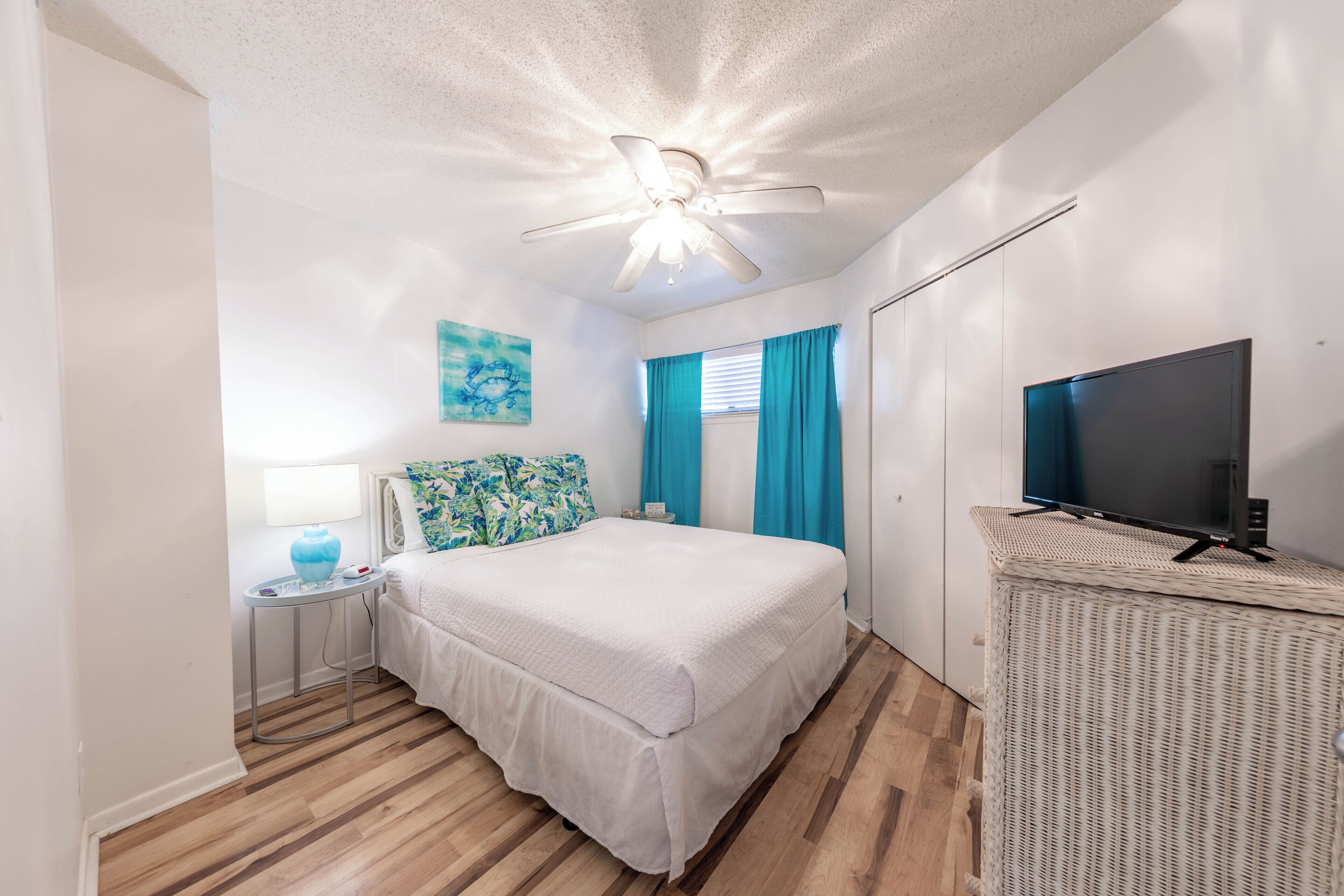 A bedroom with a bed, a television on a stand, a ceiling fan, a nightstand, a lamp, and a window with curtains.