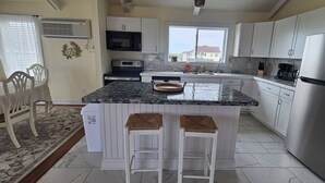 A modern kitchen with white cabinetry, a stainless steel refrigerator, a microwave, and a stove. There is a dining area with a table and chairs, and a window with blinds.