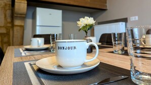 En vit mugg med texten ”BONJOUR” på ett fat, ett glas vatten och en vas med vita blommor står på ett bord i ett rum. I rummet finns ett träskåp med texten ”HEM” och ett vitt kylskåp i bakgrunden.