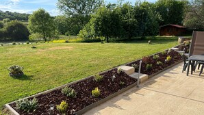 Un jardín con bancales elevados, un patio con sillas y una pequeña estructura de madera al fondo.