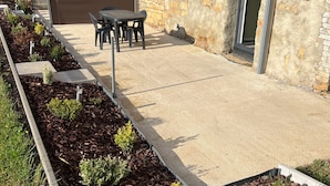 Un patio con suelo de hormigón, rodeado de bancales elevados con diversas plantas. Hay una mesa y sillas preparadas para comer al aire libre.