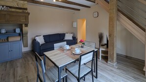 Una sala de estar acogedora con un sofá oscuro, una mesa de comedor de madera con sillas, una zona de cocina con armarios y una escalera.