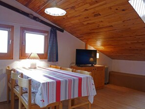 Acogedor dormitorio en el ático con techo de madera, televisor, una mesa de comedor con sillas y una ventana con cortinas.