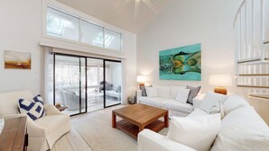 Uma sala de estar ampla com uma grande porta de vidro deslizante que dá para uma varanda, um sofá branco, uma mesa de centro de madeira e um quadro abstrato de grandes dimensões na parede.