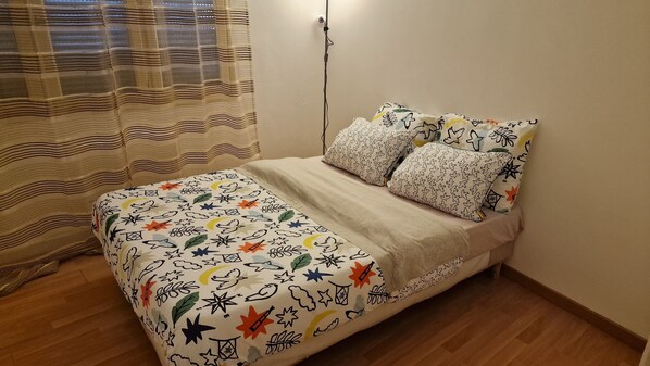A bed with a floral and star pattern bedspread, two patterned pillows, and a window with a striped curtain.