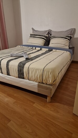 A bed with a striped bedspread and pillows, a wooden floor, and a plain wall.