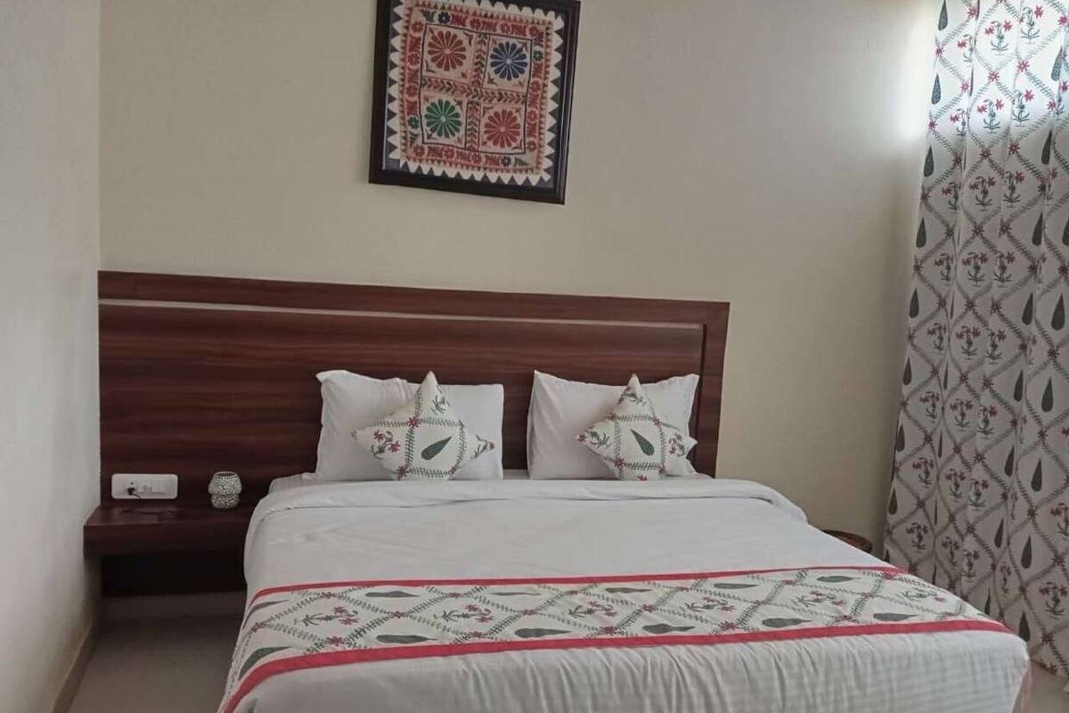 Ein ordentlich bezogenes Bett mit einem gemusterten Bettbezug und zwei Kissen mit Blumenmotiven, ein hölzernes Kopfteil, ein kleiner Nachttisch mit einer Lampe und ein gerahmtes Kunstwerk an der Wand.