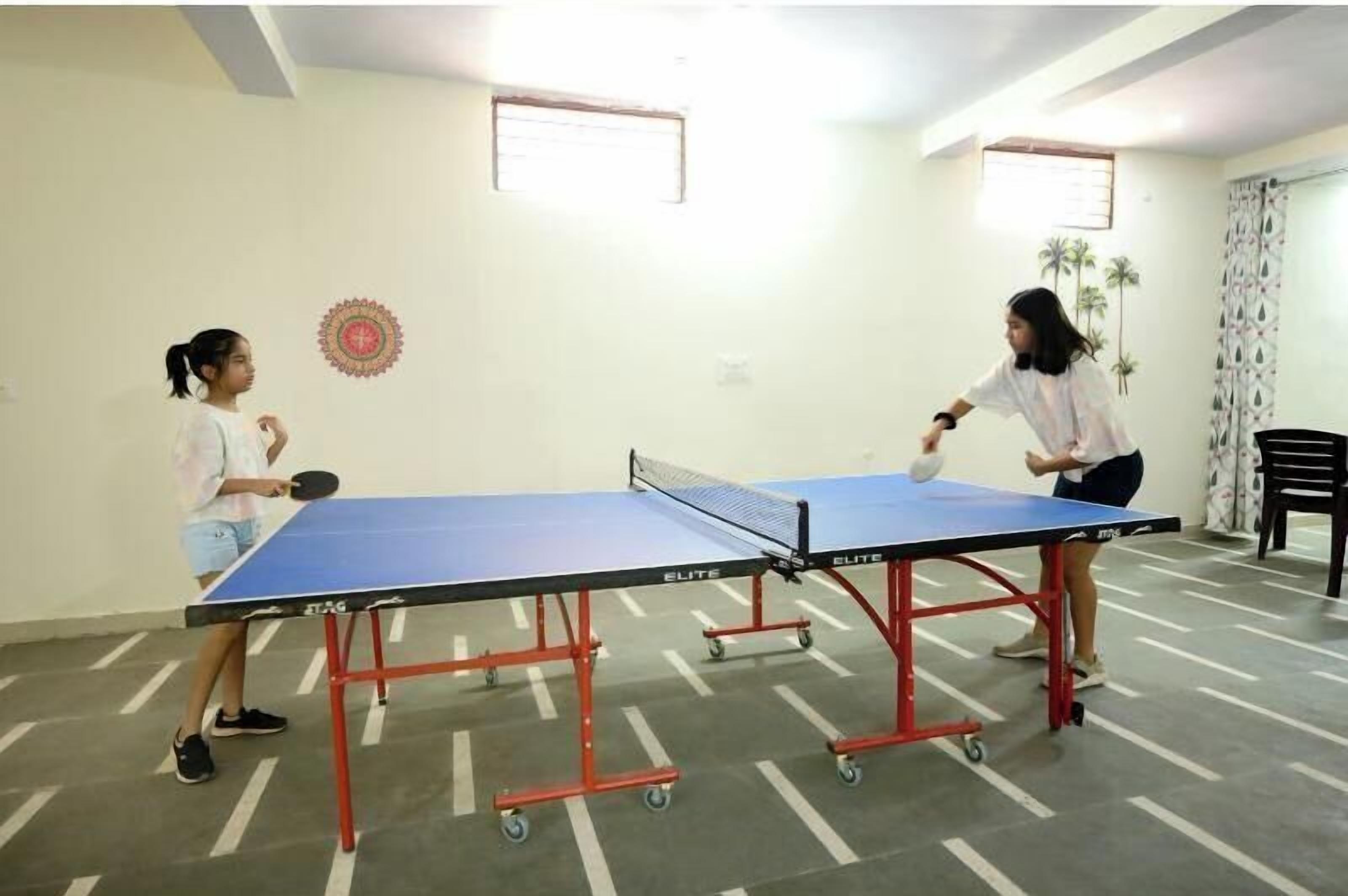 Una mesa de ping pong en una habitación con piso estampado, un reloj de pared y una ventana con cortinas.