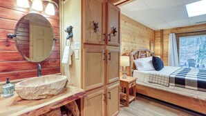 A wooden cabin interior with a stone sink, wooden cabinets, a bed with a checkered blanket, and a window with a view of trees. - Secluded Cherry Log Cabin w/ Hot Tub + Swings! (Cherry Log)
