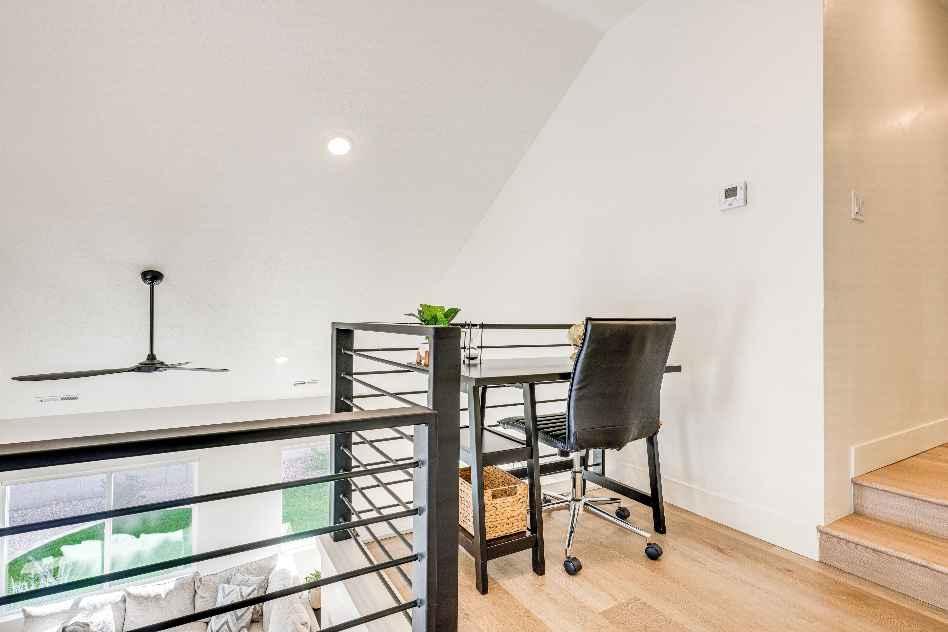 Una habitación moderna con ventilador de techo, una silla de oficina negra, una pequeña mesa y vista a un jardín a través de un ventanal grande.