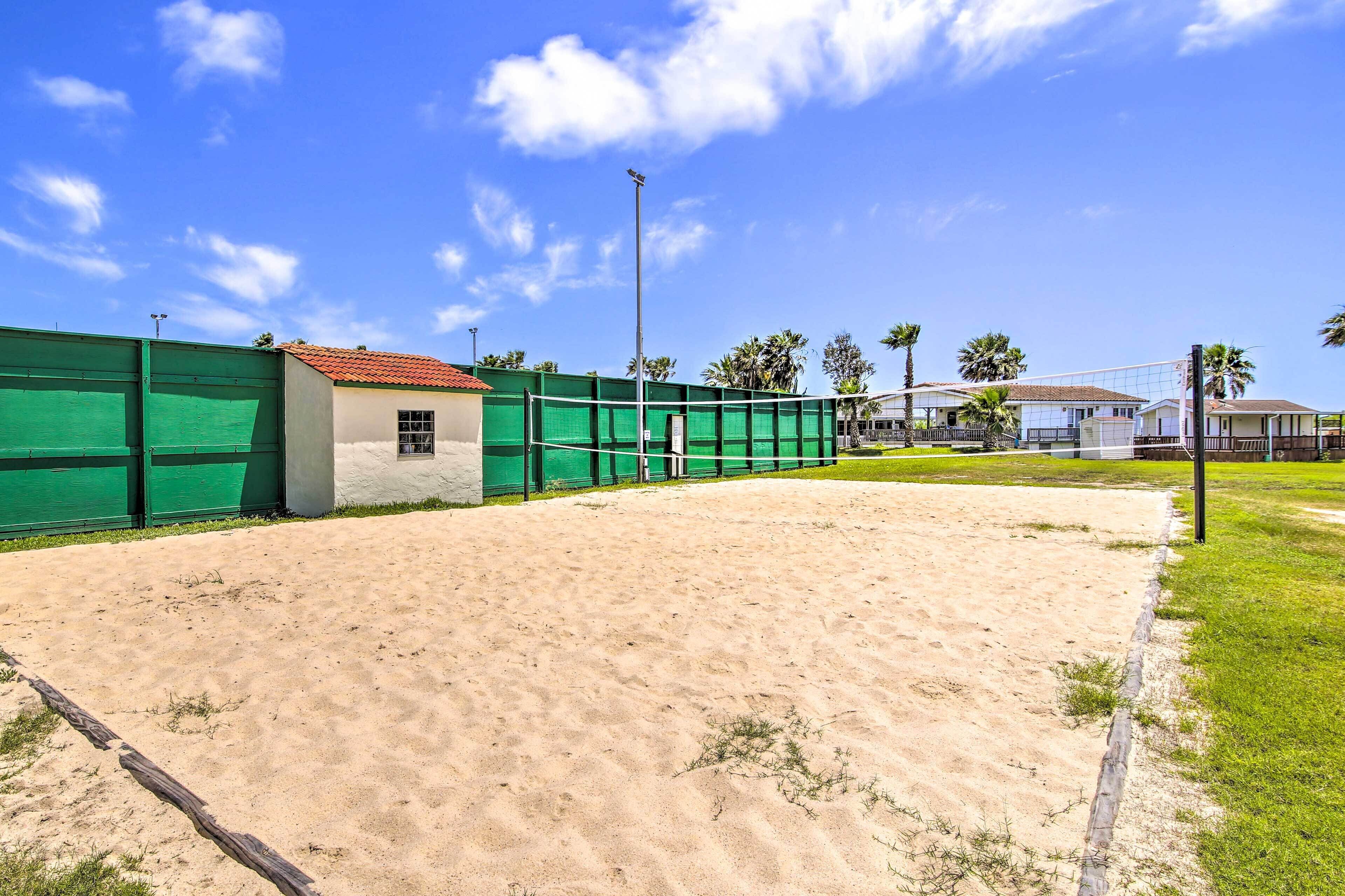A sandy volleyball court with a green fence, a small building, and palm trees in the background.