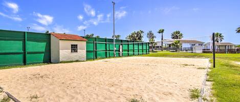 A sandy volleyball court with a green fence, a small building, and palm trees in the background.
