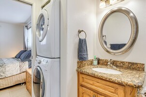 A compact hotel room with a washing machine, a bed with a patterned bedspread, a wooden vanity with a sink, and a large mirror.